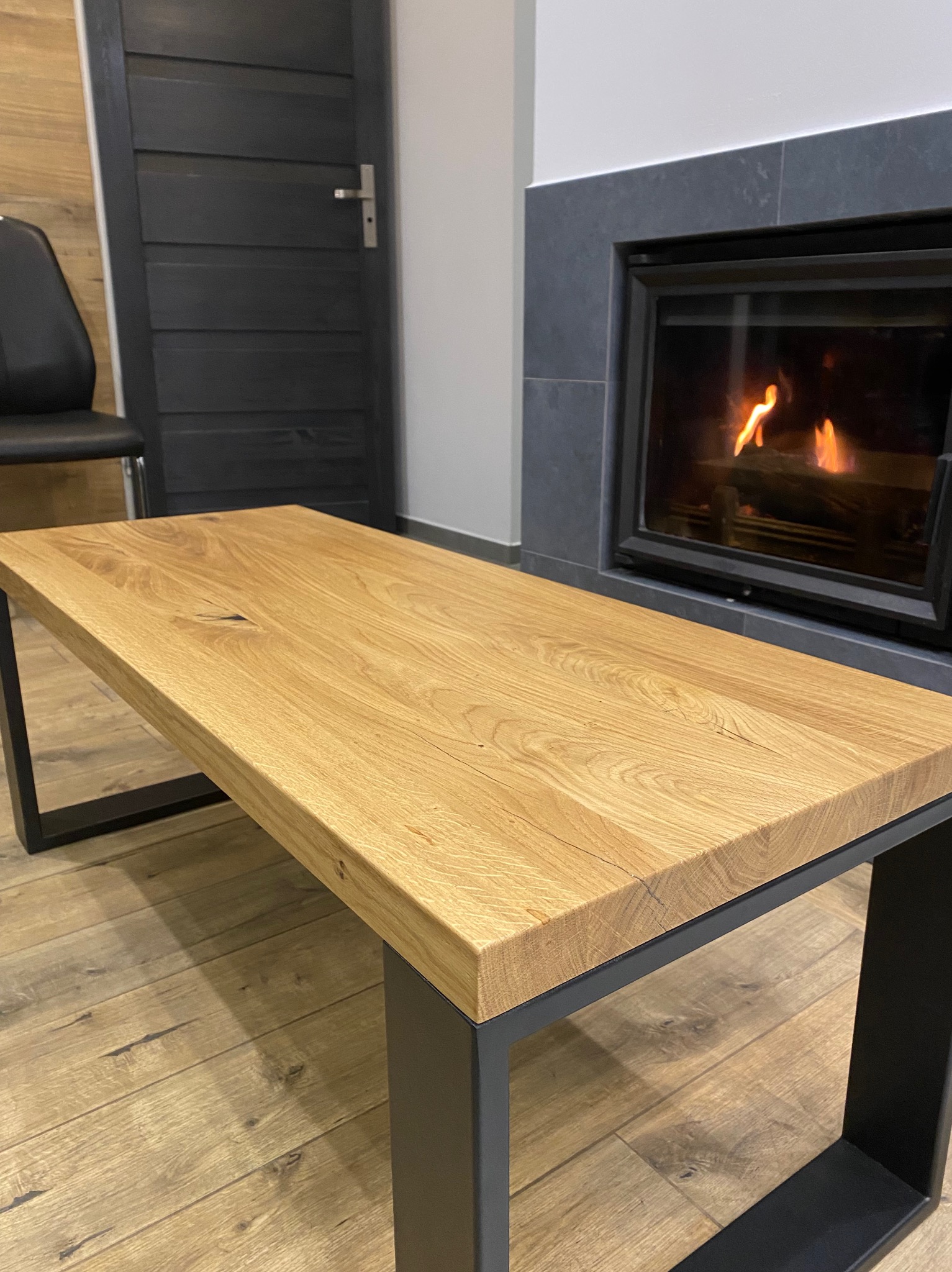 Stolik kawowy z blatem z jasnego drewna i czarnymi metalowymi nogami, ustawiony na podłodze z paneli, z kominkiem w tle.