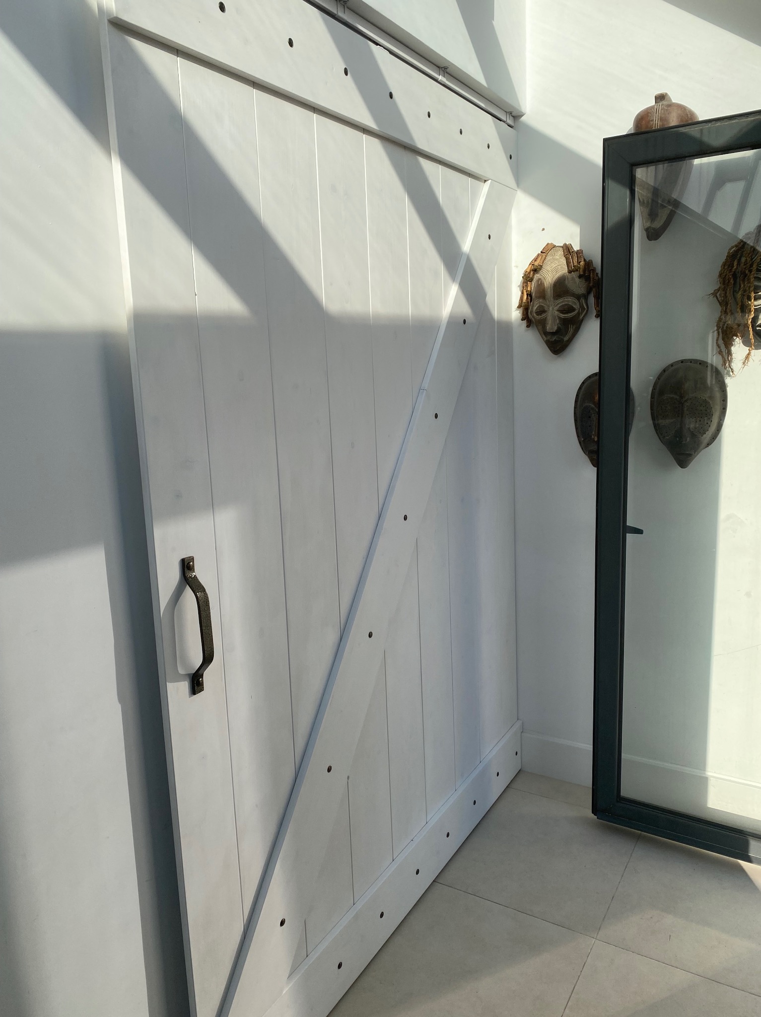 Białe, przesuwne drzwi stodołowe z metalowym uchwytem, zamontowane w nowoczesnym wnętrzu z afrykańskimi maskami w szklanej gablocie.