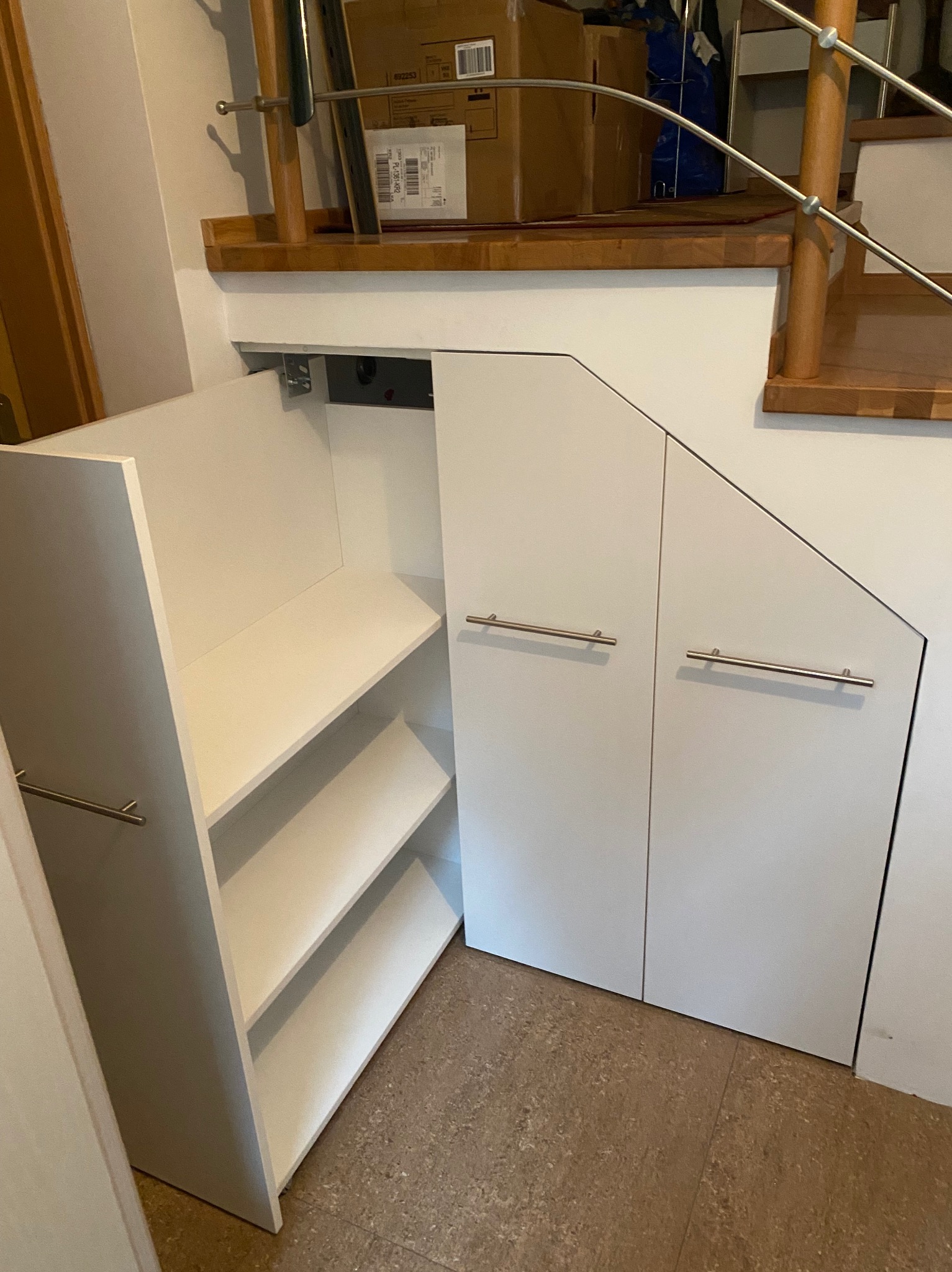 Biała zabudowa pod schodami z półkami, widoczne metalowe uchwyty, podłoga z terakoty.