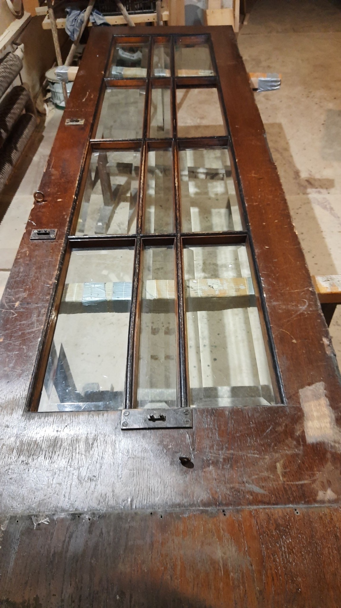 Stara drewniana rama drzwi z szybami, widoczne ślady użytkowania i niedoskonałości, leżąca poziomo na podłożu w warsztacie.