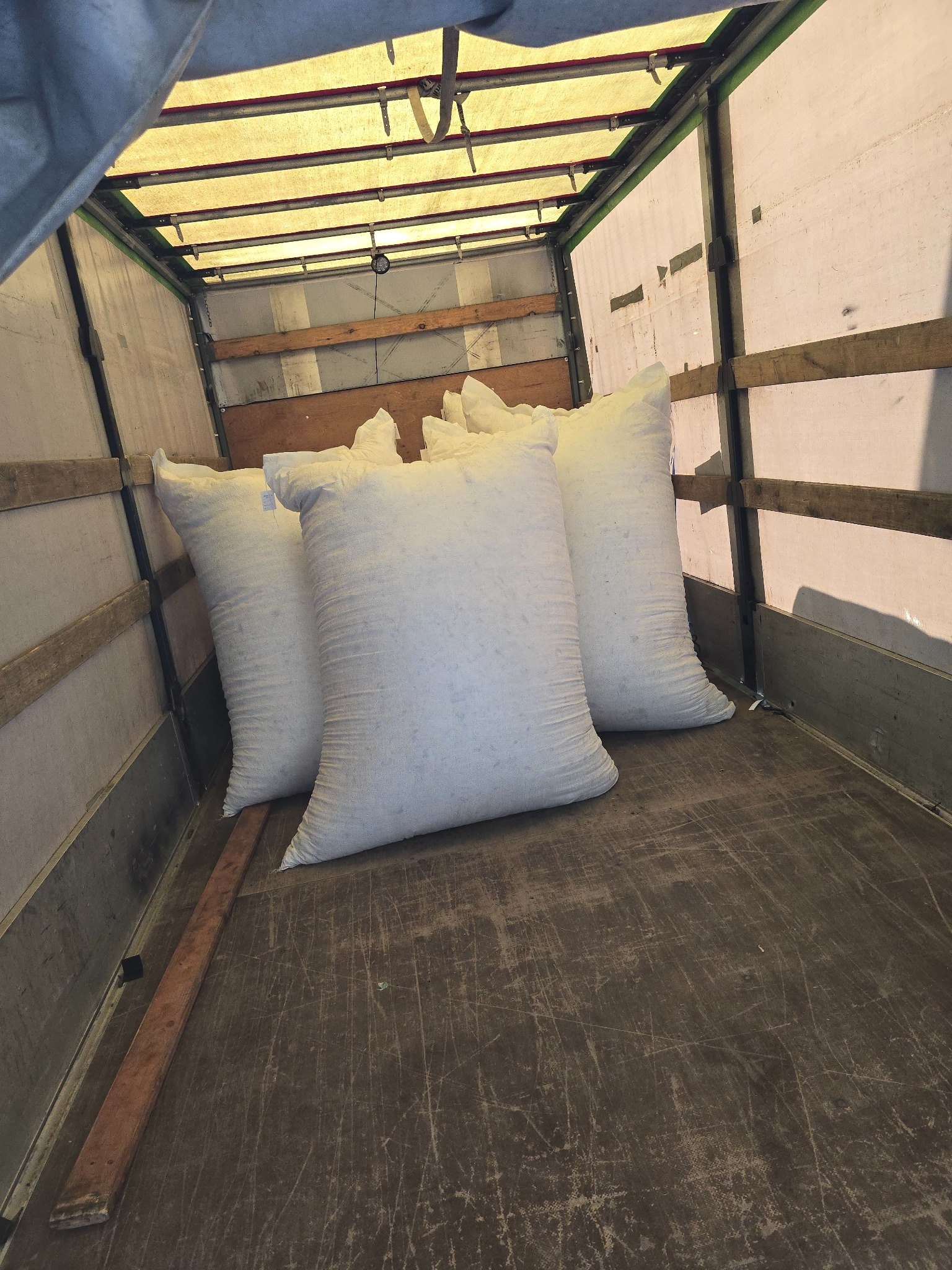 Trzy białe, pełne worki ustawione wewnątrz przestrzeni ładunkowej busa z widocznymi drewnianymi elementami podłogi i jasną plandeką na górze.