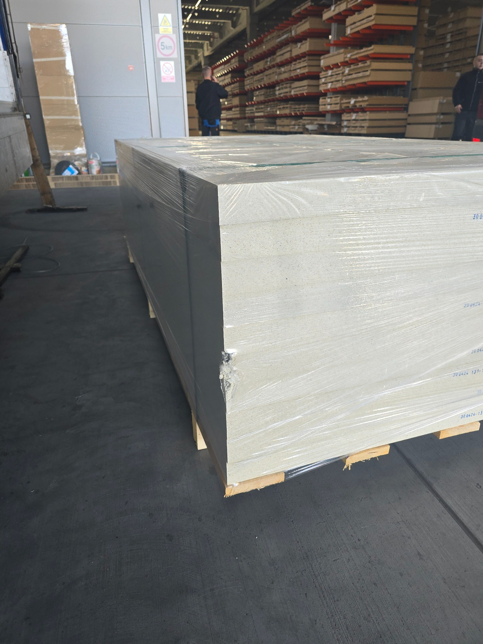 Paleta z płytami gipsowo-kartonowymi owinięta folią stretch, widoczne uszkodzenie narożnika, w tle magazyn z regałami i pracownikami.