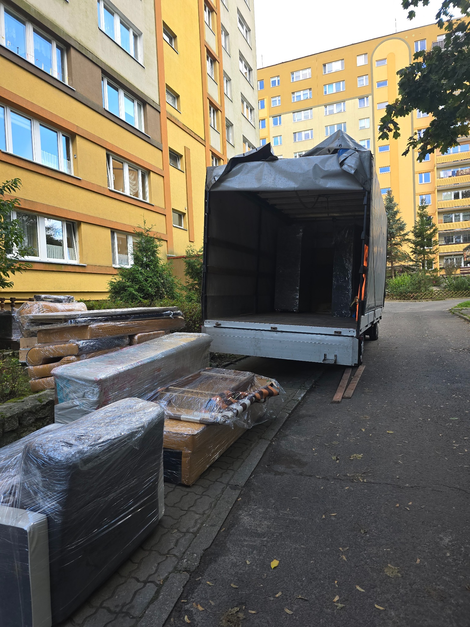Otwarta paka samochodu dostawczego na tle bloku mieszkalnego, obok leżą meble owinięte folią stretch.