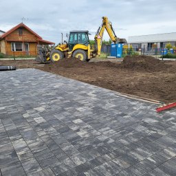 SCARAB Kamil Szubała - Ciemnoszara kostka brukowa o nieregularnym wzorze ułożona na pierwszym planie, obok przygotowany grunt z koparką New Holland w tle. Widoczne narzędzia i domy.