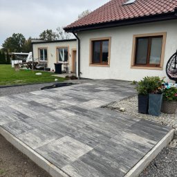 SCARAB Kamil Szubała - Nowoczesny taras z dużych, prostokątnych płyt betonowych o nieregularnym wzorze szarości, z betonowym obrzeżem, przy białym domu. Widoczne oczko wodne i trawnik.