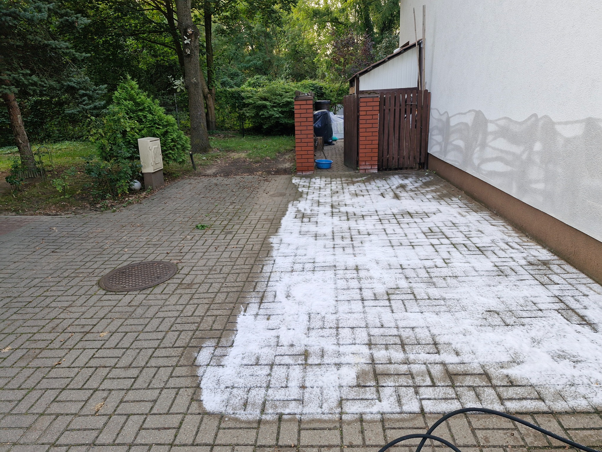 Częściowo wyczyszczona kostka brukowa na podjeździe, widoczne różnice w kolorze i zabrudzeniu, z lewej strony studzienka kanalizacyjna, w tle zieleń i fragment budynku.