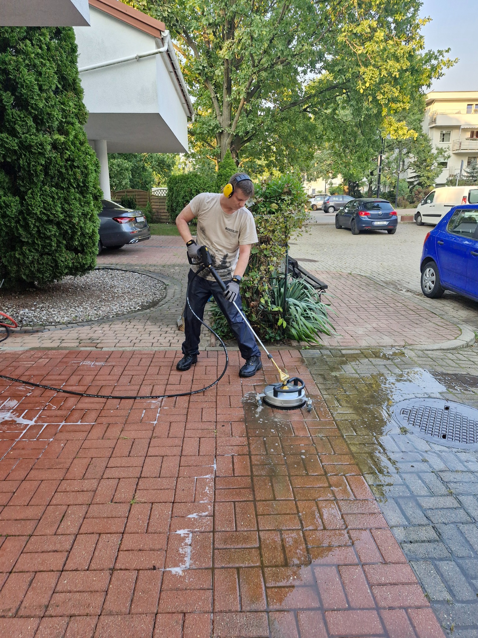 Mężczyzna w słuchawkach i rękawicach ochronnych czyści czerwoną kostkę brukową za pomocą myjki ciśnieniowej z okrągłą dyszą, widoczny efekt czyszczenia - różnica między umytą a brudną kostką.
