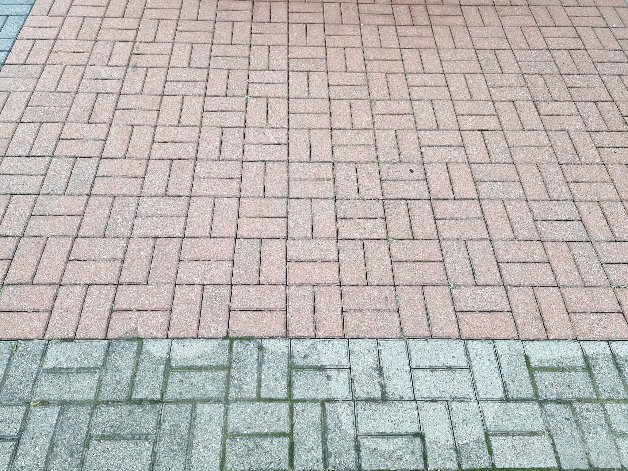 Ułożona kostka brukowa w odcieniach różu i szarości, widoczna z góry, z niewielkimi chwastami rosnącymi między niektórymi elementami.
