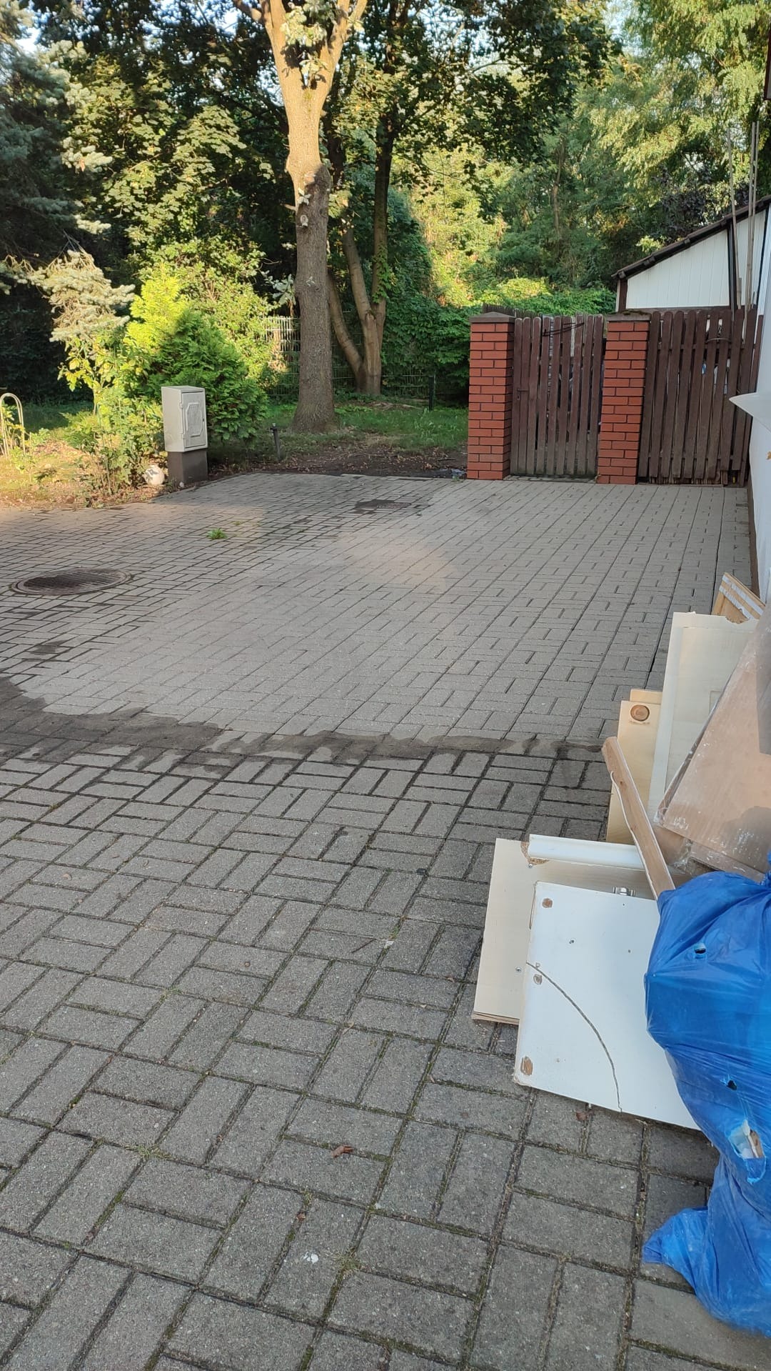 Fragment kostki brukowej z widocznymi śladami po niedawnym czyszczeniu, obok leżą śmieci i elementy mebli w niebieskim worku na tle zieleni i drzew