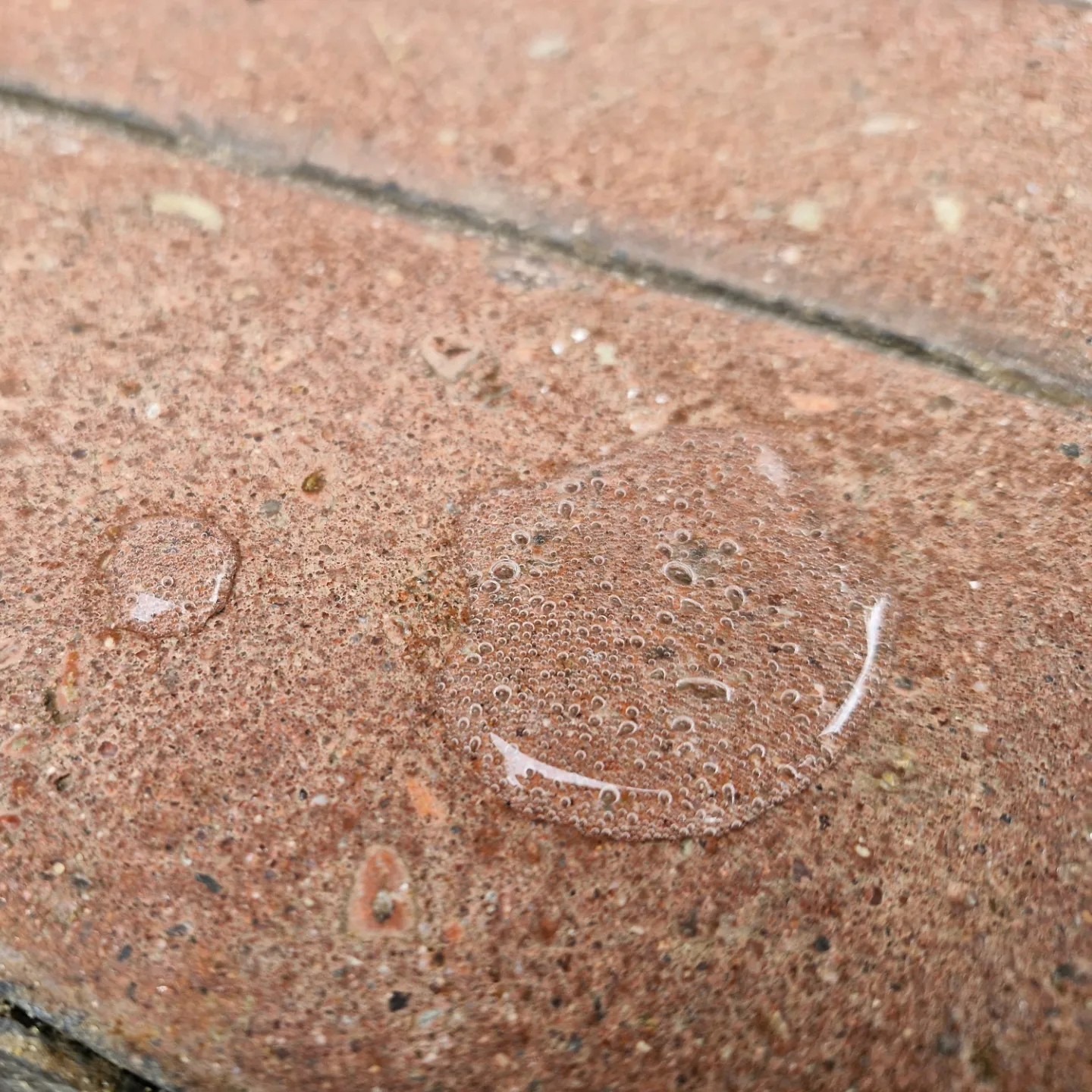 Krople wody z pęcherzykami powietrza na powierzchni czerwonej kostki brukowej, widoczne drobne nierówności i porowatość materiału.