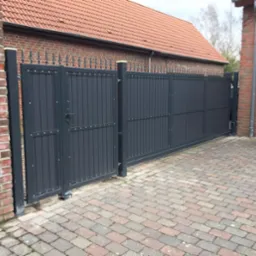 Nowoczesna, przesuwna brama wjazdowa w kolorze antracytowym, z dekoracyjnymi elementami na górze, zamontowana na posesji z ceglanym domem i brukowanym podjazdem.