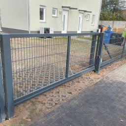 AM Metallzaune - Nowoczesne ogrodzenie panelowe w kolorze grafitowym, oddzielające posesję od chodnika z kostki brukowej, widoczne kosze na śmieci i elementy wyposażenia ogrodu w tle.