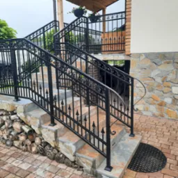 Zewnętrzne schody z czarną, metalową balustradą o zdobieniach w kształcie grotów, prowadzące do wejścia z kamienną elewacją i drewnianym zadaszeniem, z ułożonym drewnem pod schodami.