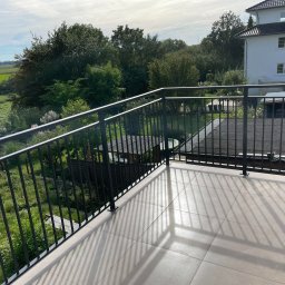 AM Metallzaune - Nowoczesny balkon z czarną metalową balustradą, jasnymi płytkami podłogowymi i widokiem na zielony ogród oraz okoliczne drzewa.