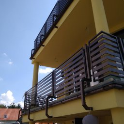 AM Metallzaune - Dwukondygnacyjny budynek z żółtą elewacją i balkonami z poziomymi, ciemnymi balustradami, widok z dołu na błękitne niebo z chmurami.