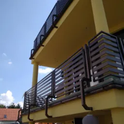 Dwukondygnacyjny budynek z żółtą elewacją i balkonami z poziomymi, ciemnymi balustradami, widok z dołu na błękitne niebo z chmurami.