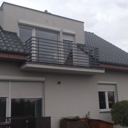 AM Metallzaune - Balkon z metalową balustradą o prostej konstrukcji, widok z dołu na szary budynek z roletami zewnętrznymi, pochmurne niebo w tle.