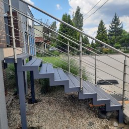 AM Metallzaune - Zewnętrzne schody z szarego metalu z balustradą ze stali nierdzewnej, prowadzące do domu z cegły, otoczone kamieniami i zielenią.