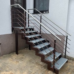 AM Metallzaune - Zewnętrzne schody z metalową konstrukcją w kolorze brązowym i srebrnymi poręczami, prowadzące do ciemnych drzwi wejściowych w białym budynku z widocznym oknem z firanką.