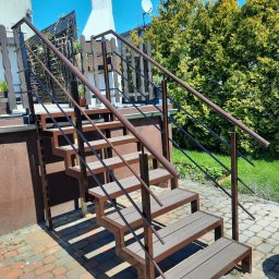 AM Metallzaune - Zewnętrzne schody z metalową konstrukcją i kompozytowymi stopniami prowadzące na taras, z czarnymi poręczami, na tle ceglanego chodnika i zieleni.