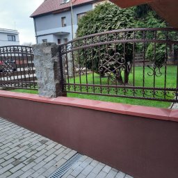 AM Metallzaune - Kute ogrodzenie z ozdobnymi elementami w kolorze ciemnego brązu, osadzone na niskim murku z kamiennymi słupkami, widok z chodnika z kostki brukowej, w tle budynki mieszkalne i zieleń.