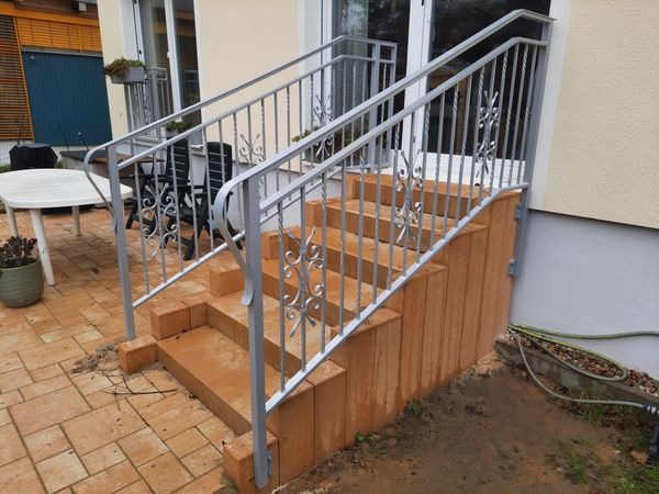 Zewnętrzne schody z jasnego kamienia z metalową balustradą o dekoracyjnych elementach, prowadzące do drzwi tarasowych.