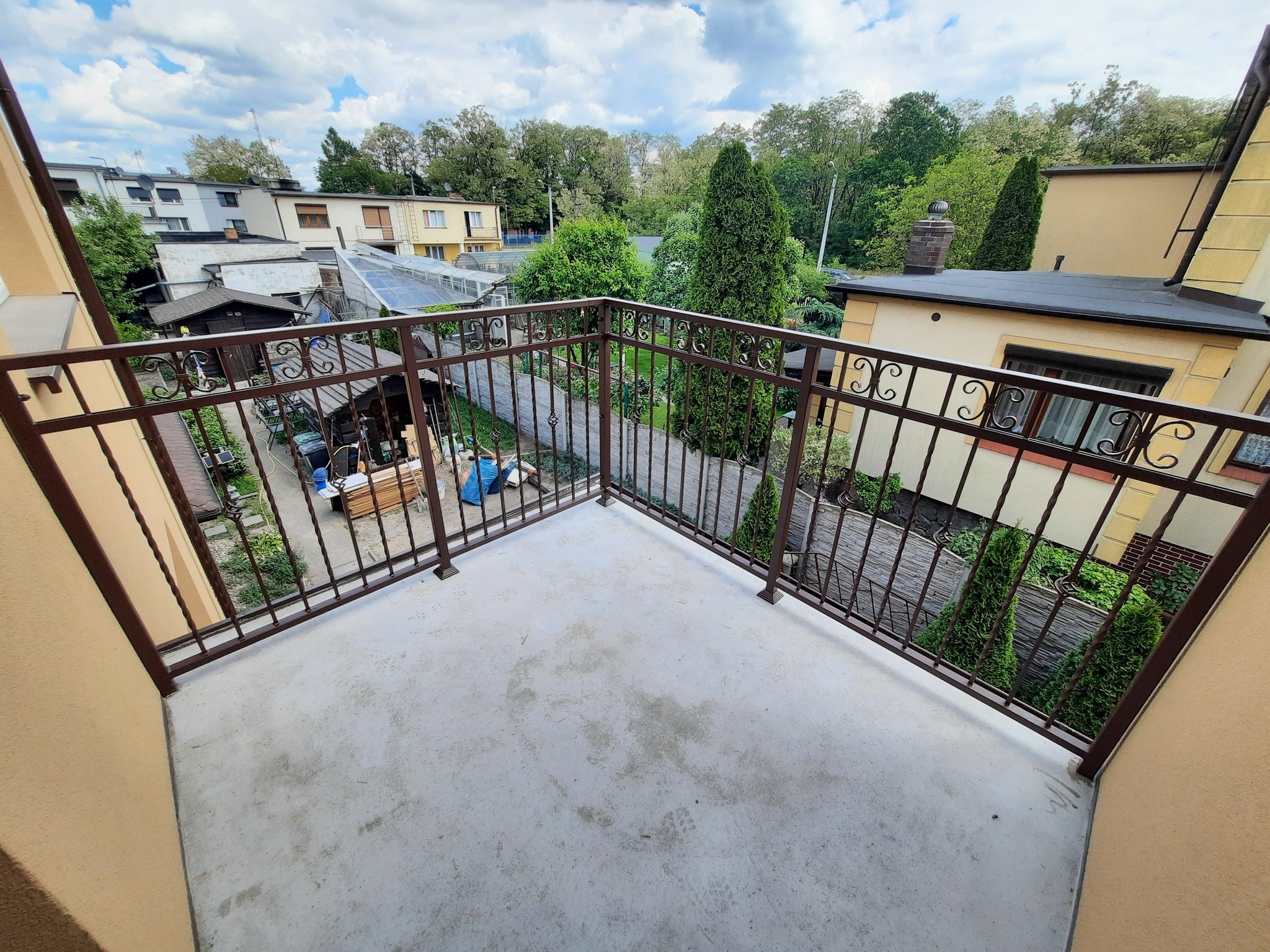 Balkon z brązową, metalową balustradą z widokiem na podwórze z zielenią i zabudowaniami w tle, uchwycony z góry.