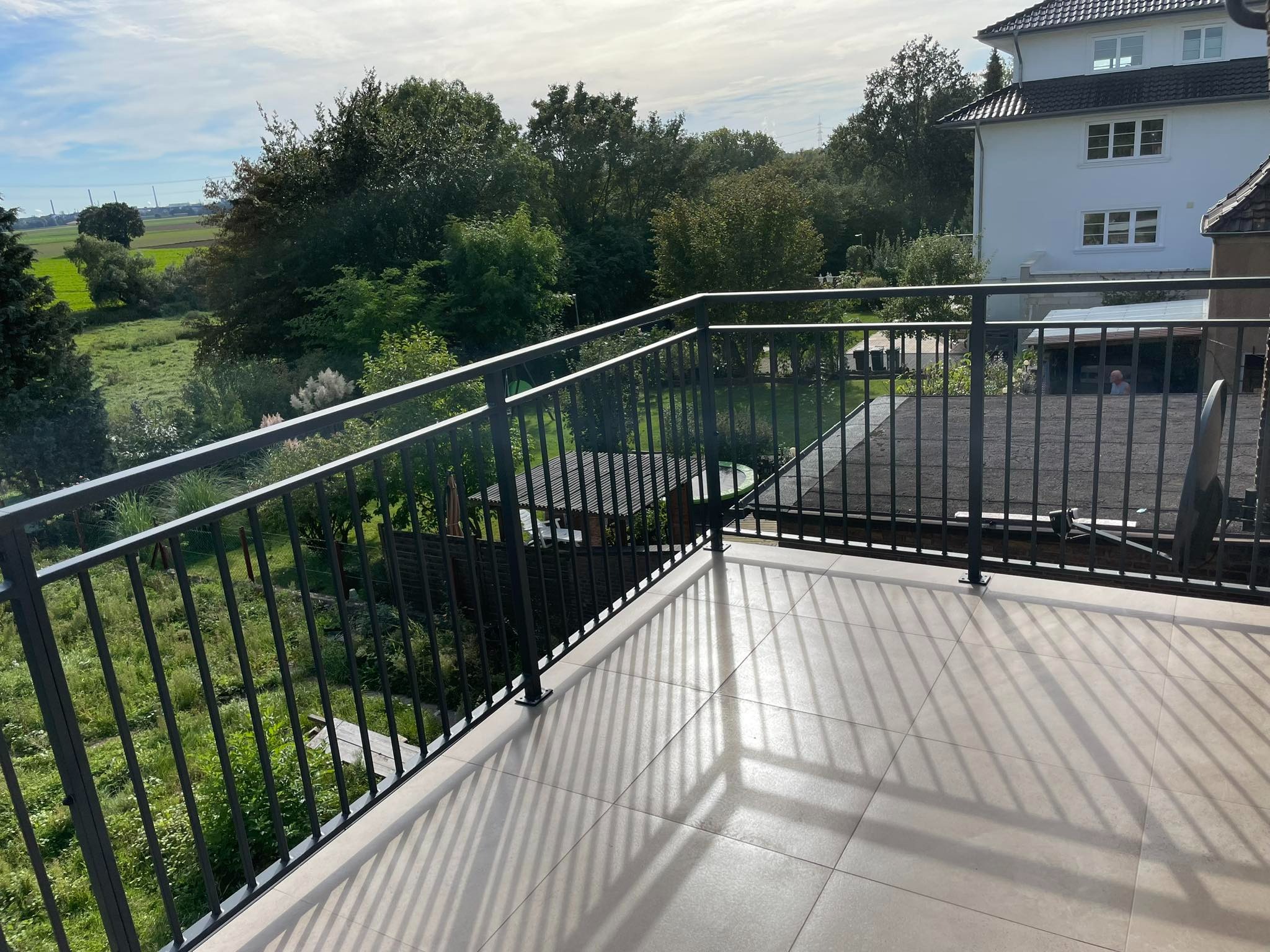 Nowoczesny balkon z czarną metalową balustradą, jasnymi płytkami podłogowymi i widokiem na zielony ogród oraz okoliczne drzewa.