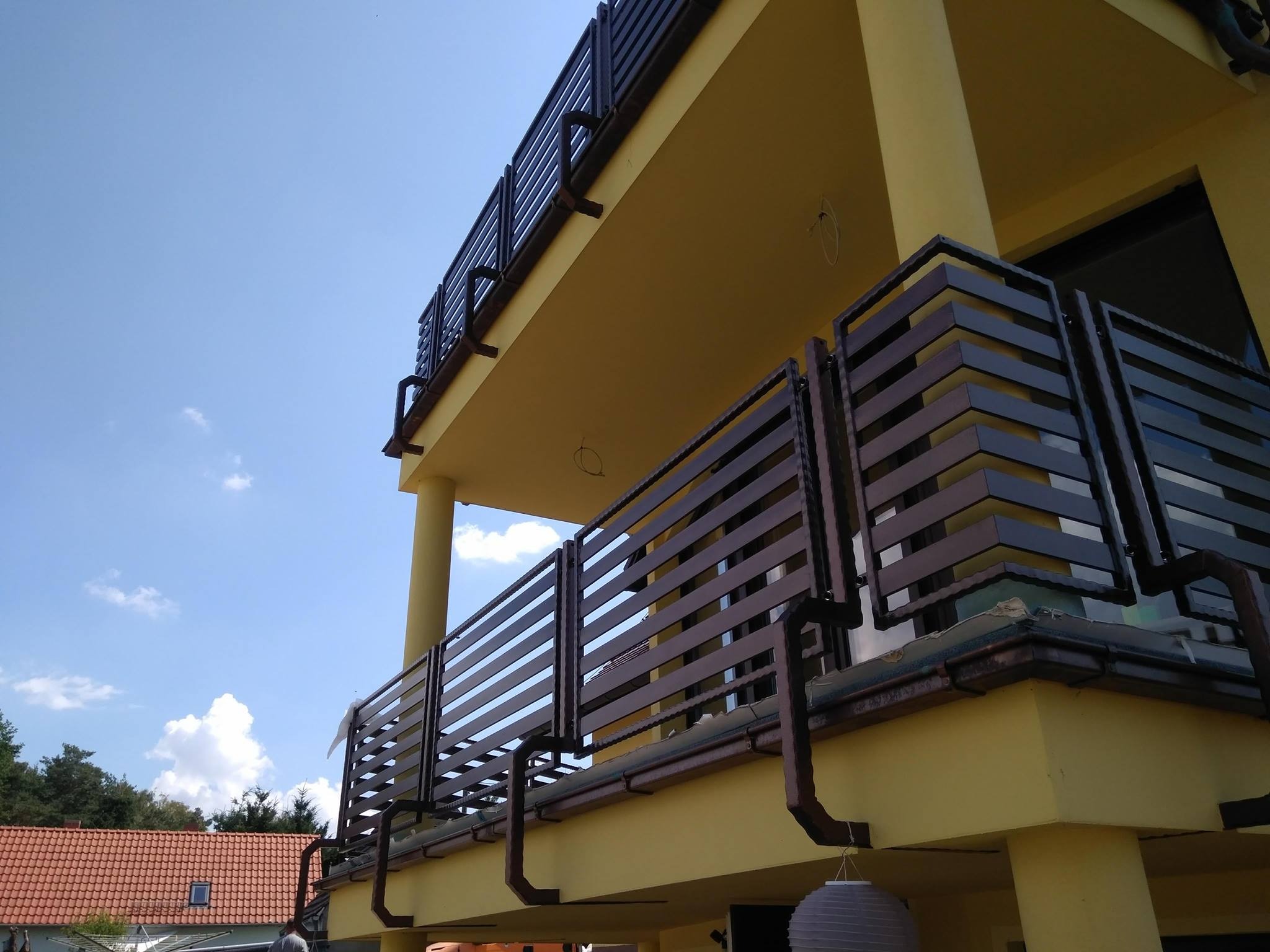 Dwukondygnacyjny budynek z żółtą elewacją i balkonami z poziomymi, ciemnymi balustradami, widok z dołu na błękitne niebo z chmurami.