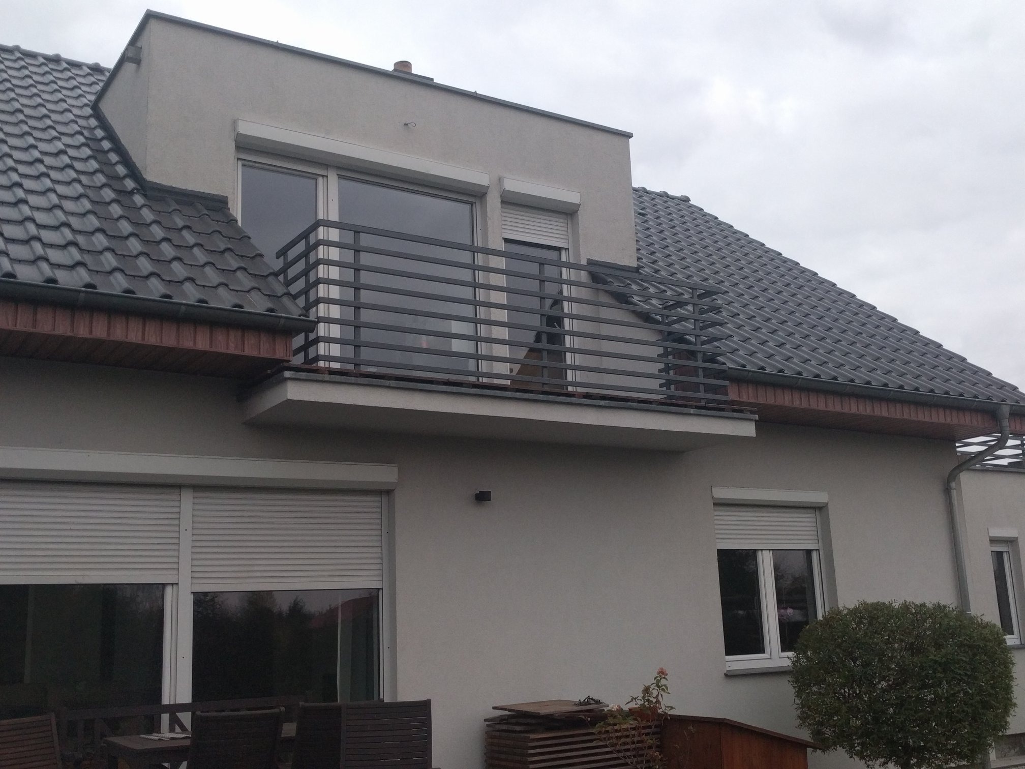 Balkon z metalową balustradą o prostej konstrukcji, widok z dołu na szary budynek z roletami zewnętrznymi, pochmurne niebo w tle.