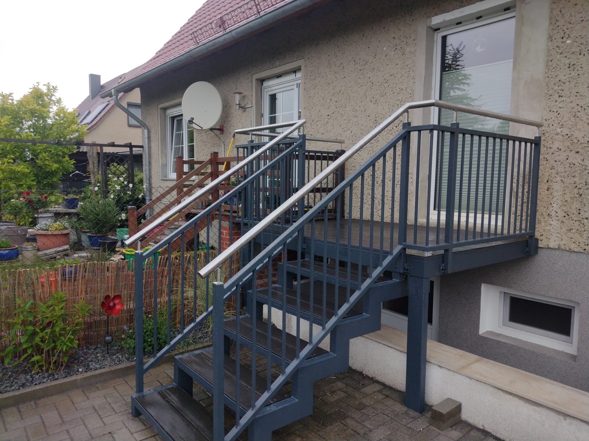 Zewnętrzne schody metalowe z drewnianymi stopniami i stalowymi poręczami prowadzące do drzwi na podwyższeniu, na tle elewacji budynku z widocznym ogrodem.