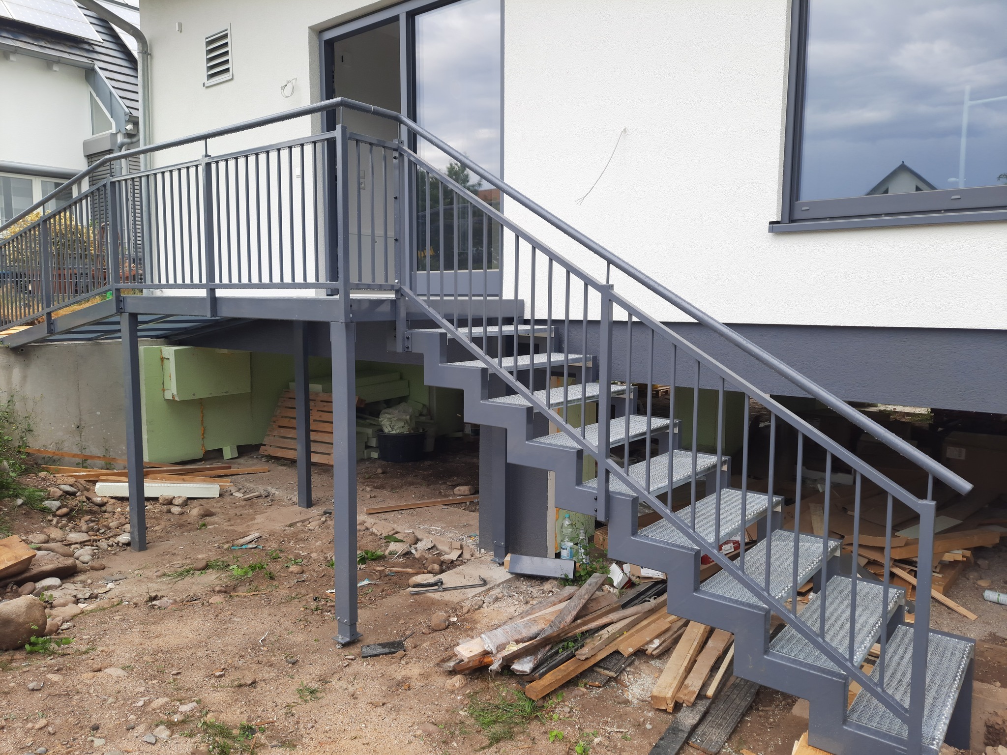 Zewnętrzne metalowe schody z ażurowymi stopniami i szarą balustradą prowadzące do przeszklonych drzwi tarasowych, widoczne ślady prac budowlanych wokół.