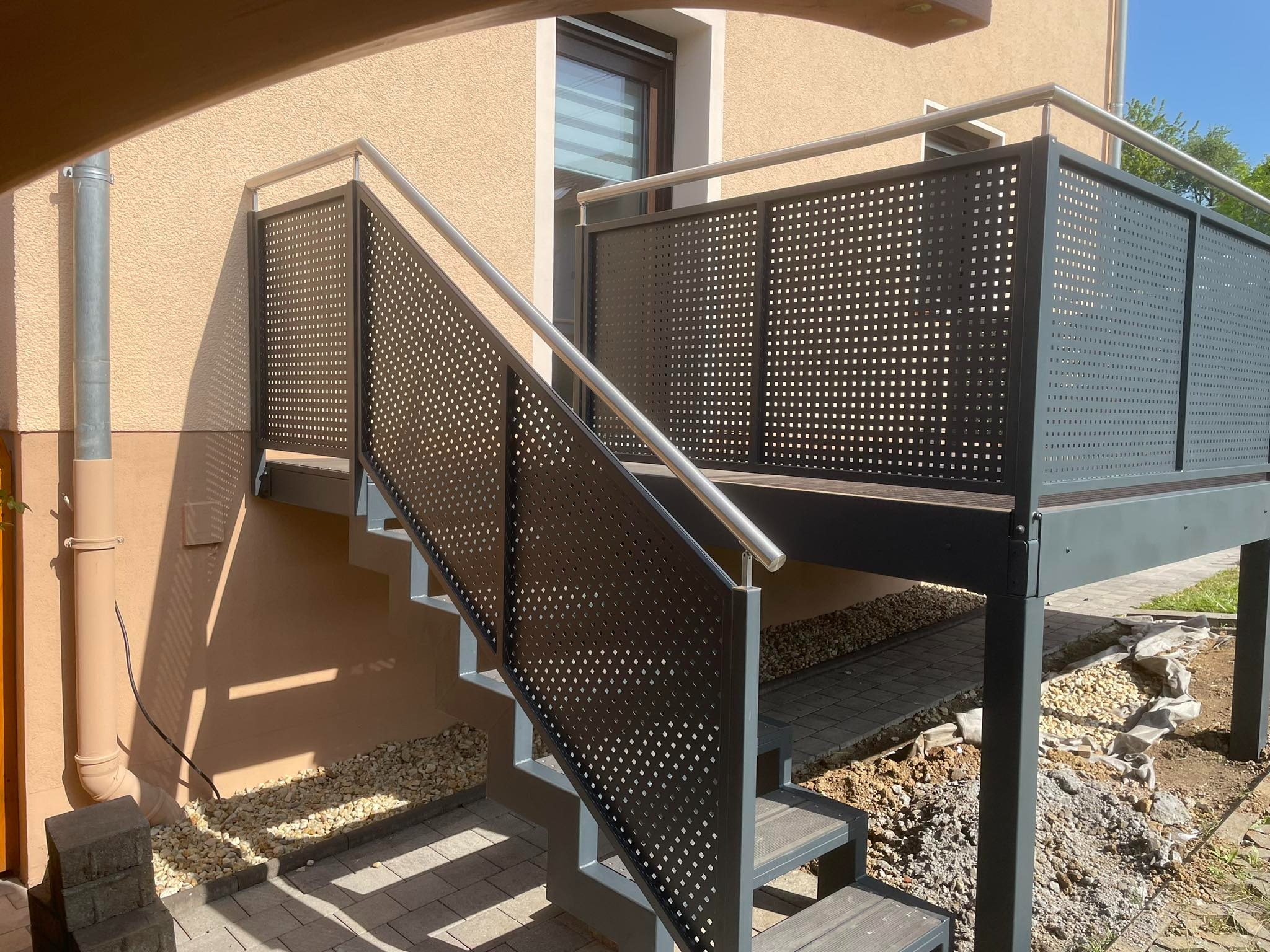 Zewnętrzne schody z metalową perforowaną balustradą i poręczą ze stali nierdzewnej, prowadzące na podest z podobną balustradą, widok na elewację budynku z oknem.