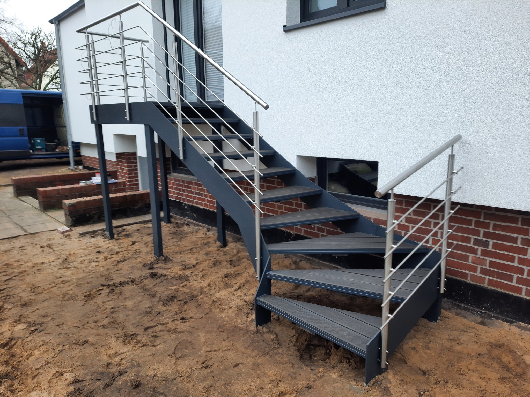 Zewnętrzne schody metalowe z szarymi stopniami kompozytowymi i nierdzewną balustradą, prowadzące do białego budynku z ceglanym fundamentem, teren wokół w trakcie prac ziemnych.