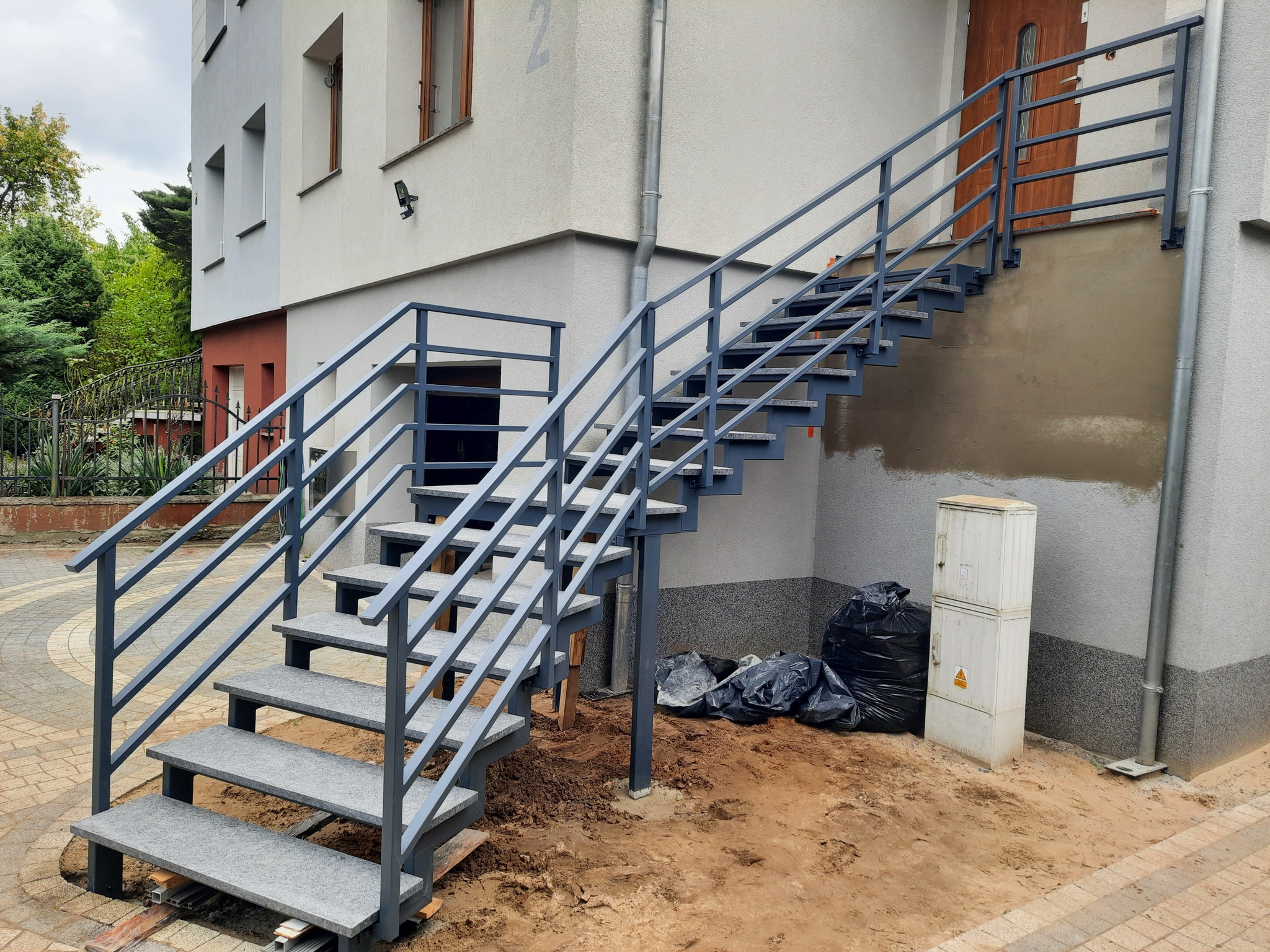 Zewnętrzne schody metalowe z szarymi stopniami prowadzące do wejścia budynku, z widocznymi śladami prac ziemnych wokół podstawy schodów i workami na śmieci w tle.