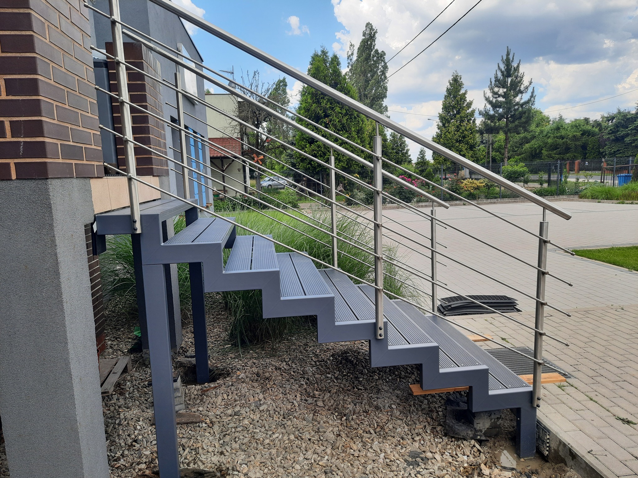 Zewnętrzne schody z szarego metalu z balustradą ze stali nierdzewnej, prowadzące do domu z cegły, otoczone kamieniami i zielenią.