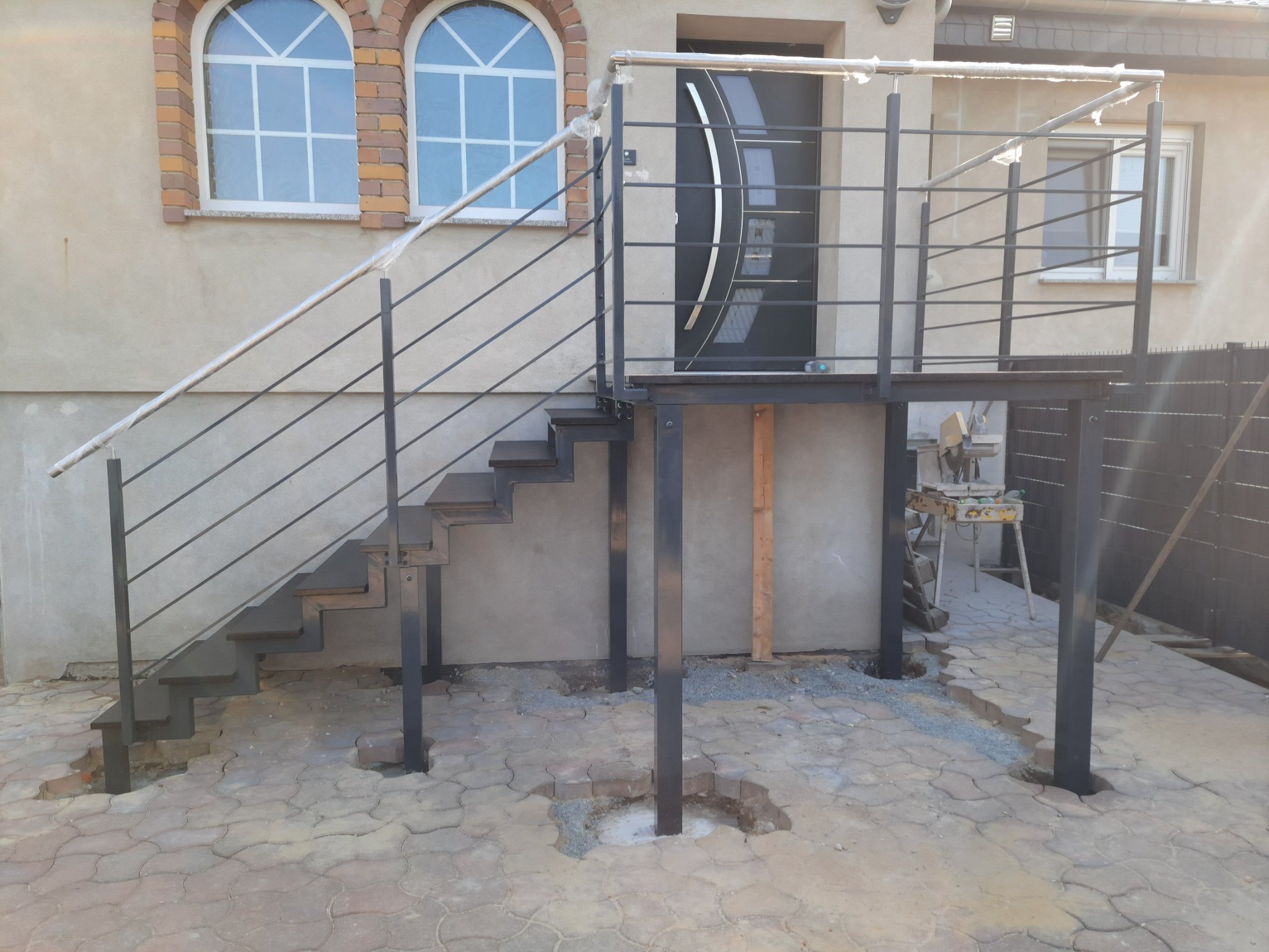 Montaż zewnętrznych schodów metalowych z poręczą, widoczne niedokończone prace brukarskie wokół słupków, zabezpieczona folią poręcz.