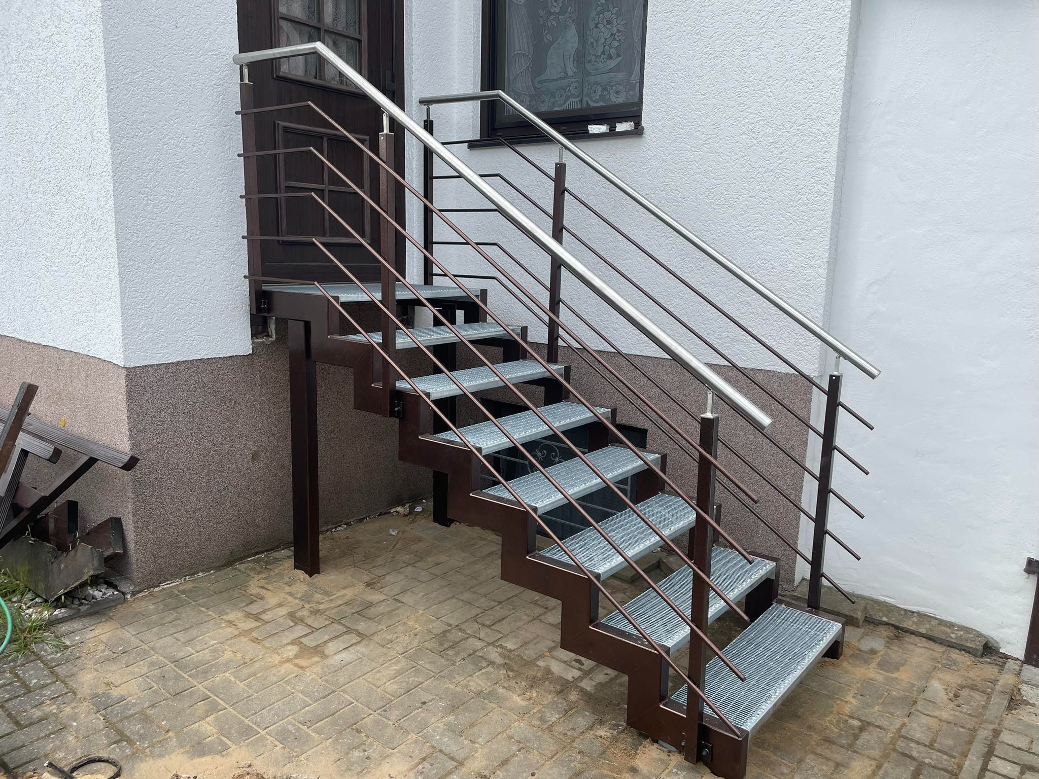 Zewnętrzne schody z metalową konstrukcją w kolorze brązowym i srebrnymi poręczami, prowadzące do ciemnych drzwi wejściowych w białym budynku z widocznym oknem z firanką.