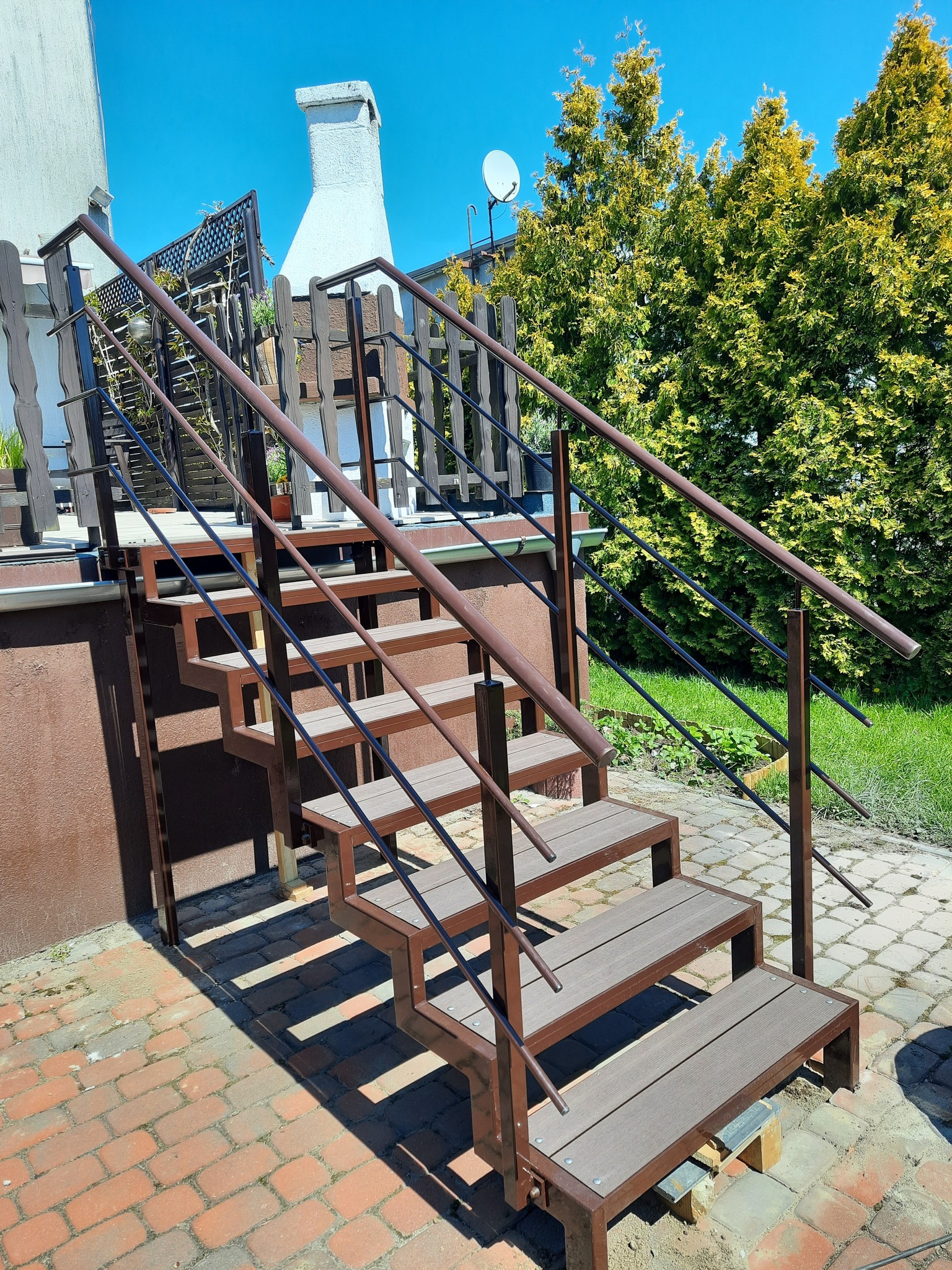 Zewnętrzne schody z metalową konstrukcją i kompozytowymi stopniami prowadzące na taras, z czarnymi poręczami, na tle ceglanego chodnika i zieleni.