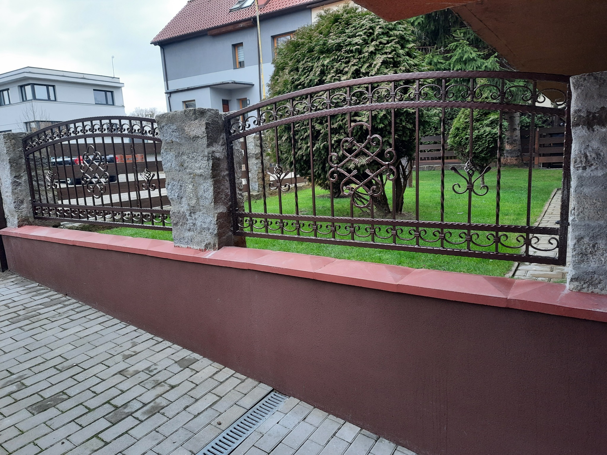 Kute ogrodzenie z ozdobnymi elementami w kolorze ciemnego brązu, osadzone na niskim murku z kamiennymi słupkami, widok z chodnika z kostki brukowej, w tle budynki mieszkalne i zieleń.