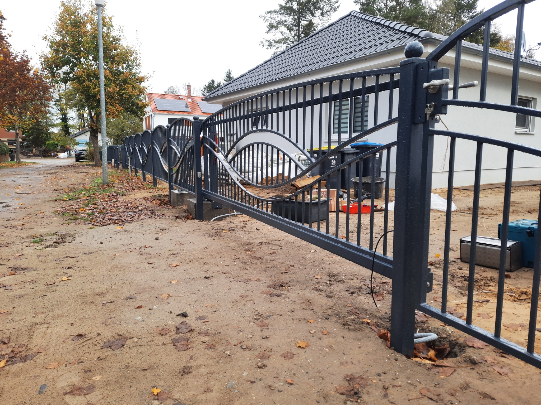 Czarna metalowa brama wjazdowa z dekoracyjnymi, falistymi elementami, częściowo otwarta, widoczne narzędzia i materiały budowlane, w tle dom z szarą dachówką i drzewa z jesiennymi liśćmi.