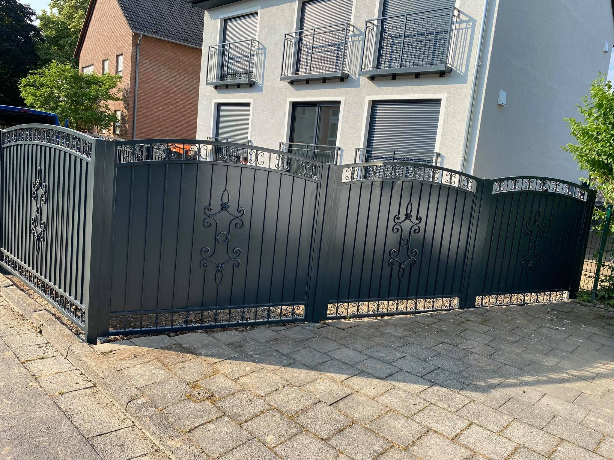 Czarna, metalowa brama wjazdowa z ozdobnymi elementami, prowadząca do domu z szarą elewacją i balkonami z metalowymi balustradami.