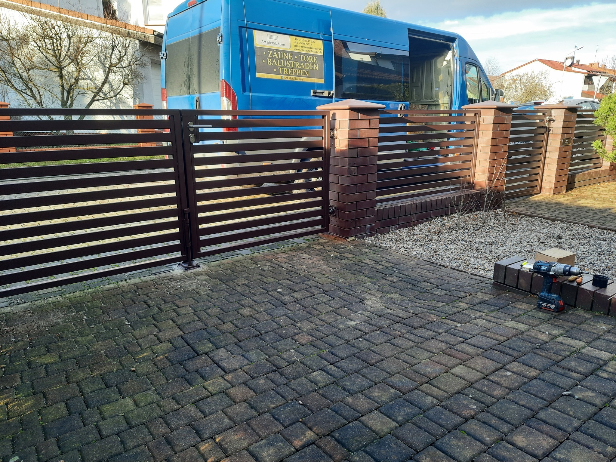 Nowoczesna brama wjazdowa z poziomych profili w kolorze brązowym, osadzona na ceglanych słupkach, z niebieskim busem firmy w tle i wiertarką akumulatorową na kostce brukowej w trakcie montażu.