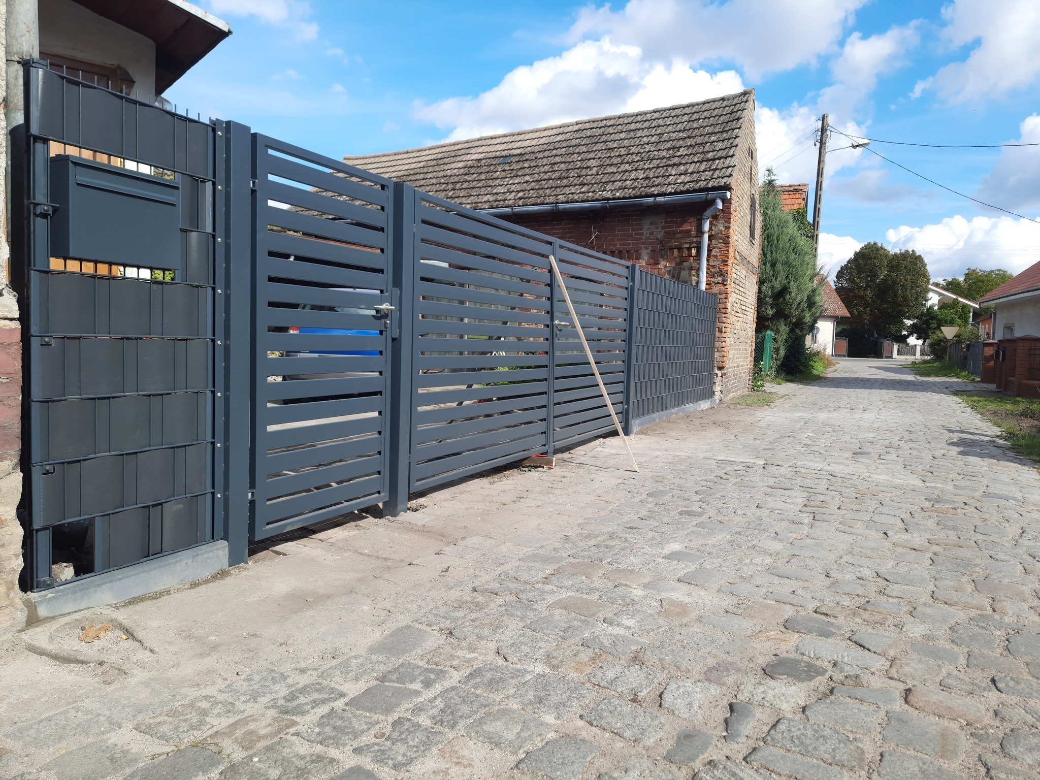 Nowoczesna, pozioma brama wjazdowa z szarego metalu z zamontowaną skrzynką na listy, widoczna z zewnątrz posesji, na tle brukowanej drogi i zabudowań.