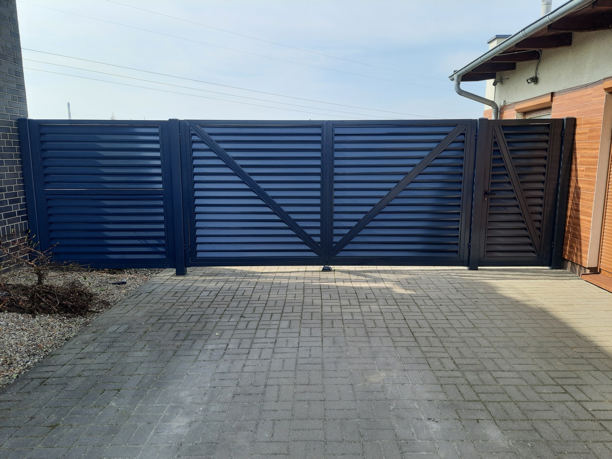 Nowoczesna, pozioma brama wjazdowa w kolorze antracytowym, z ukośnymi wzmocnieniami, zamontowana na posesji z brukowanym podjazdem i widocznym fragmentem domu.
