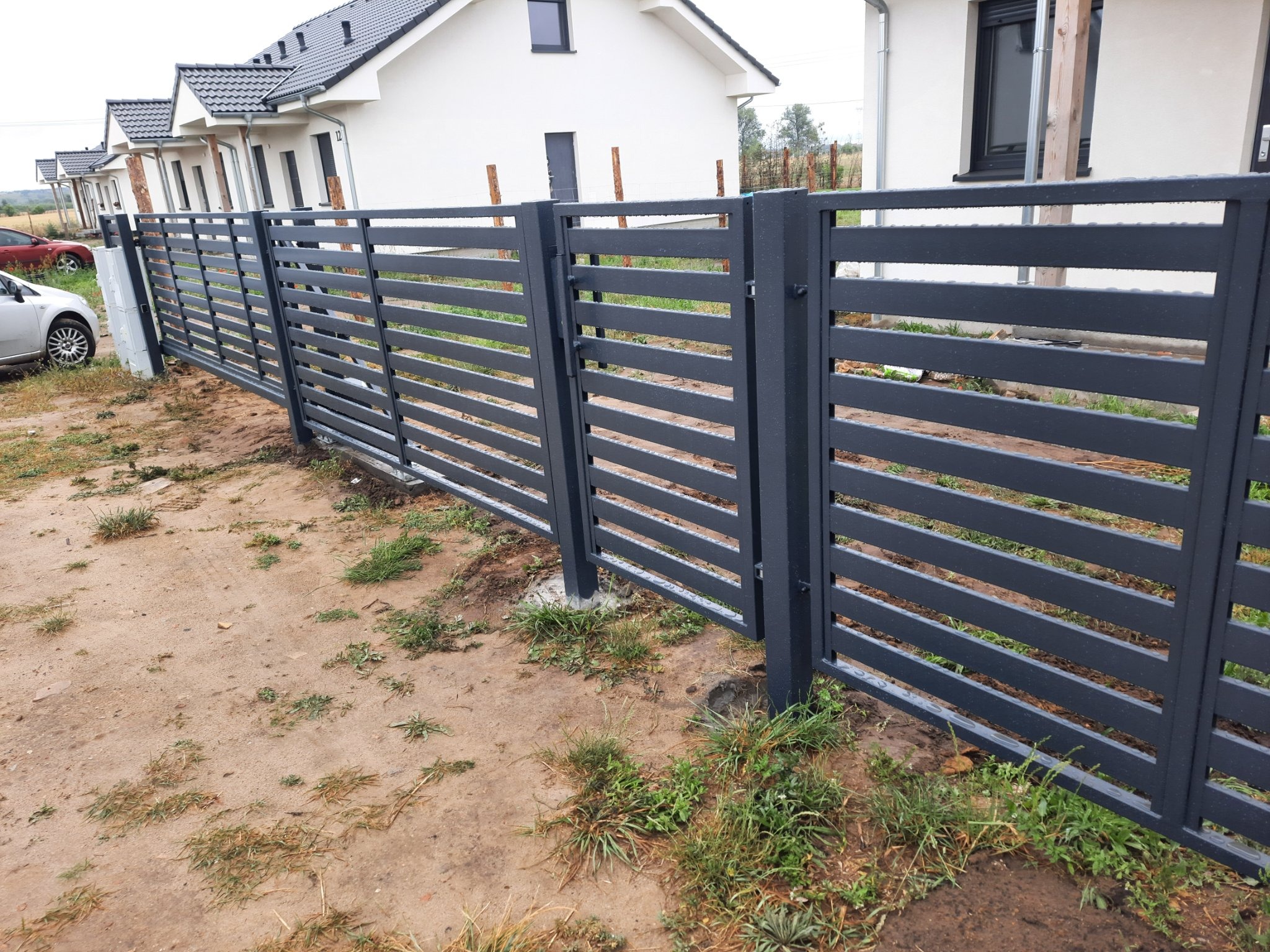 Nowoczesne, poziome ogrodzenie metalowe w kolorze grafitowym, otaczające posesję z nowymi domami jednorodzinnymi w tle, widoczne ślady niedawnych prac ziemnych.