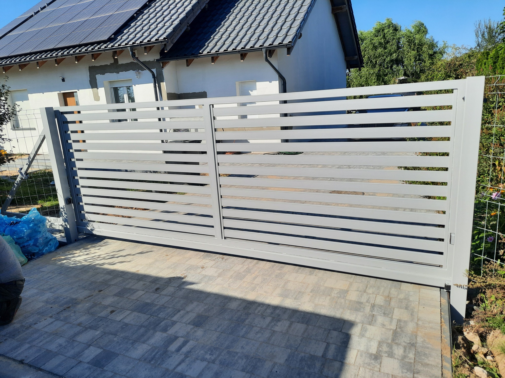 Nowoczesna brama wjazdowa przesuwna, wykonana z poziomych, szarych profili metalowych, na tle domu z panelami słonecznymi i kostki brukowej.