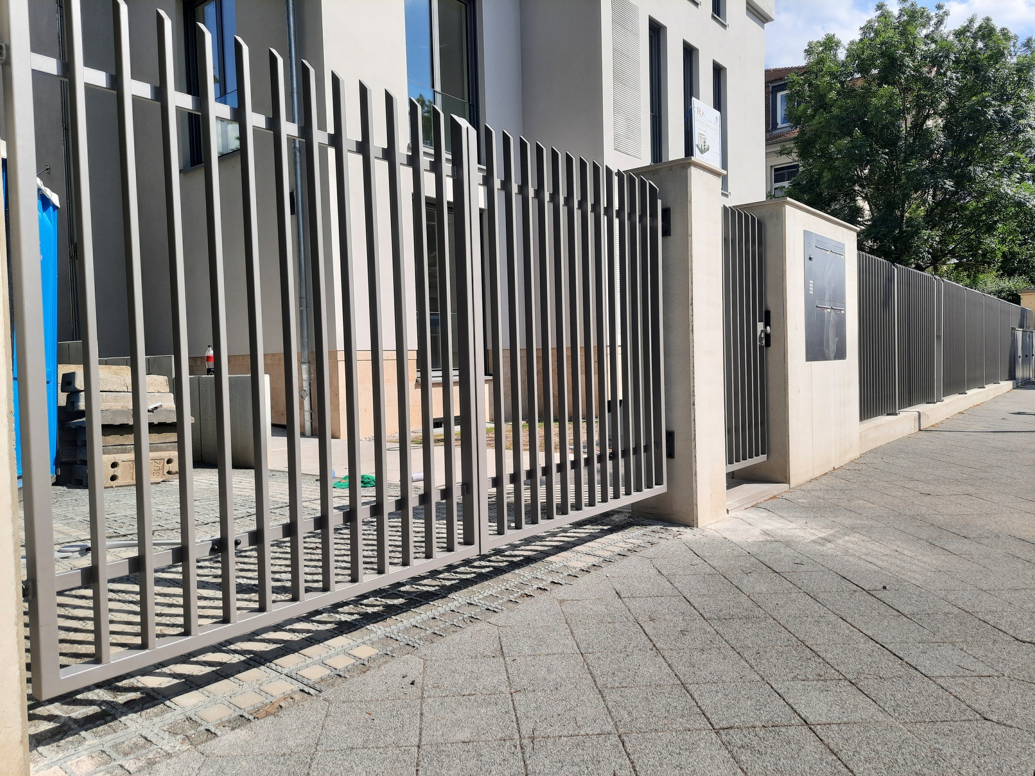 Nowoczesna, szara brama wjazdowa z pionowymi szczeblami, zintegrowana z ogrodzeniem posesji, na tle jasnego budynku i chodnika z kostki.