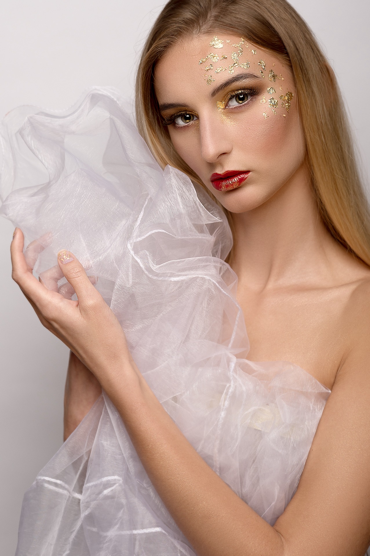 Portret kobiety z blond włosami, z intensywnym złotym zdobieniem na twarzy w postaci płatków i błyszczącej szminki, owiniętej w białą, prześwitującą tkaninę.