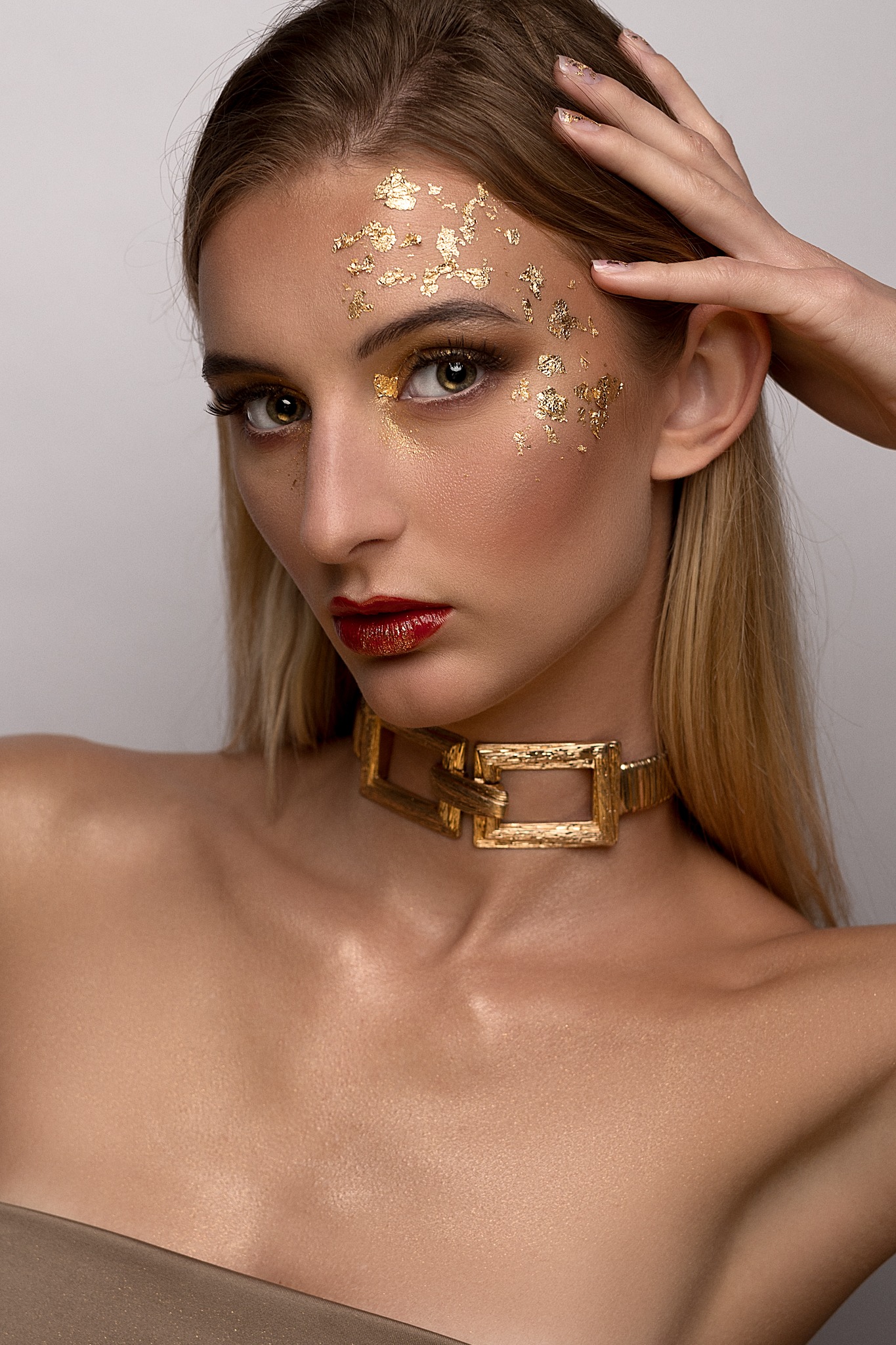Portret kobiety z artystycznym zdobieniem twarzy złotymi płatkami, intensywnie czerwone usta i złota biżuteria na szyi.